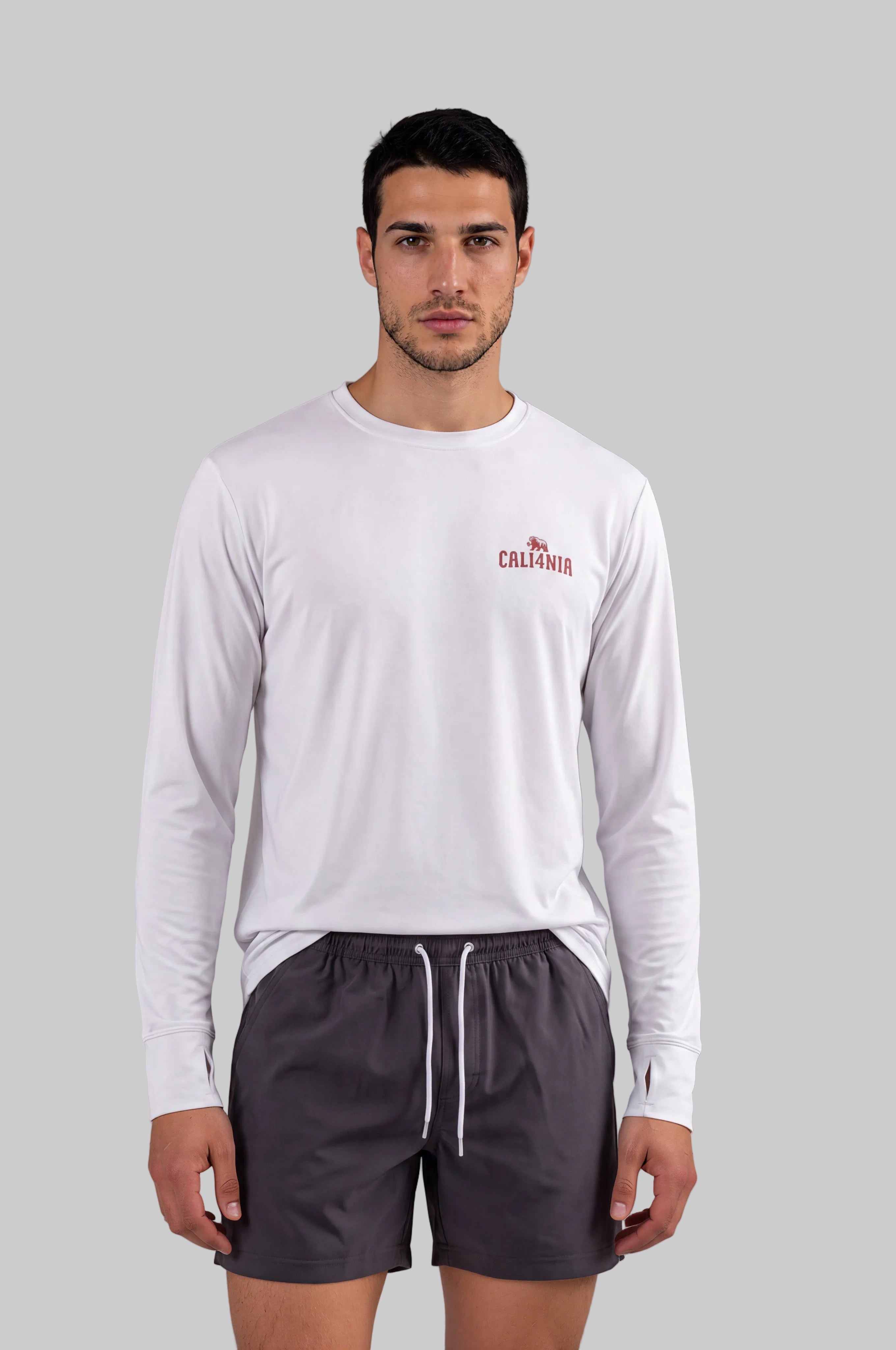 A man modeling a white long-sleeve crewneck shirt with a small red 'CALIFORNIA' logo on the left chest, paired with dark gray athletic shorts, against a solid gray studio background.