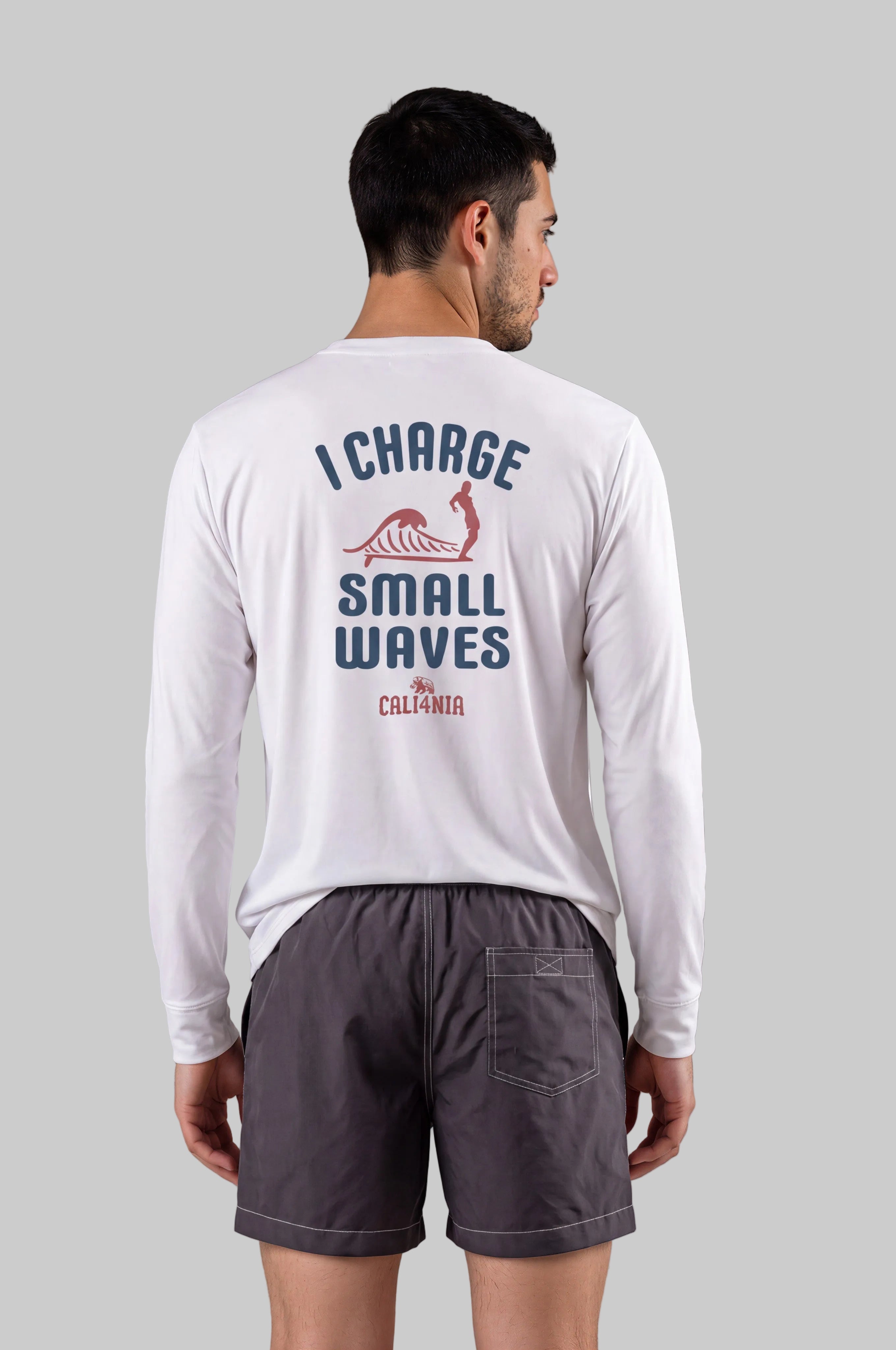 A man shown from the back wearing a white long-sleeve t-shirt with a blue and red graphic on the back that reads 'I CHARGE SMALL WAVES CALIFORNIA' and dark gray shorts, against a solid gray studio background.