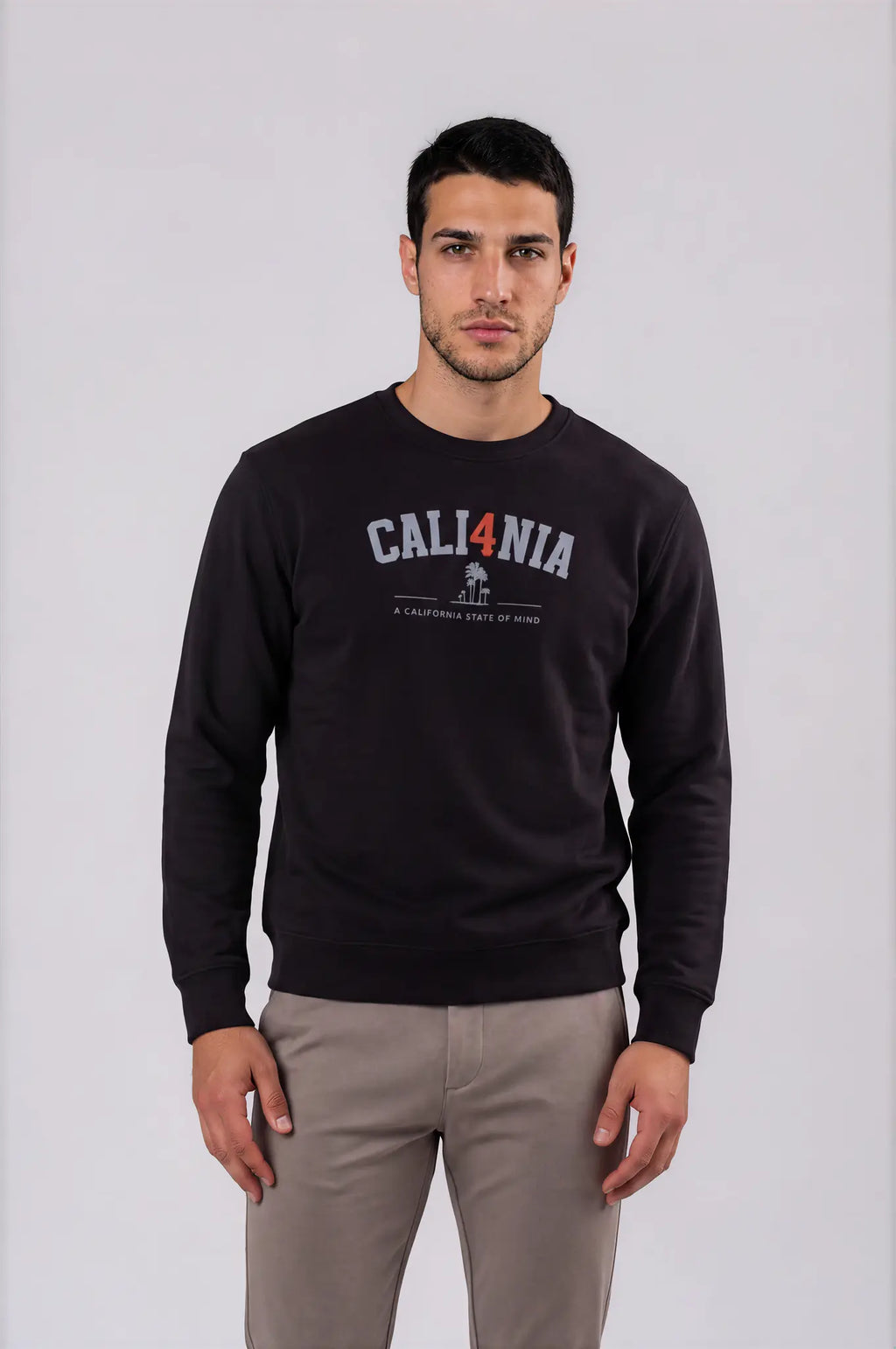 A man wearing a black crewneck sweatshirt with a 'CALI4NIA' graphic on the chest, paired with light gray trousers, standing against a light gray studio background.