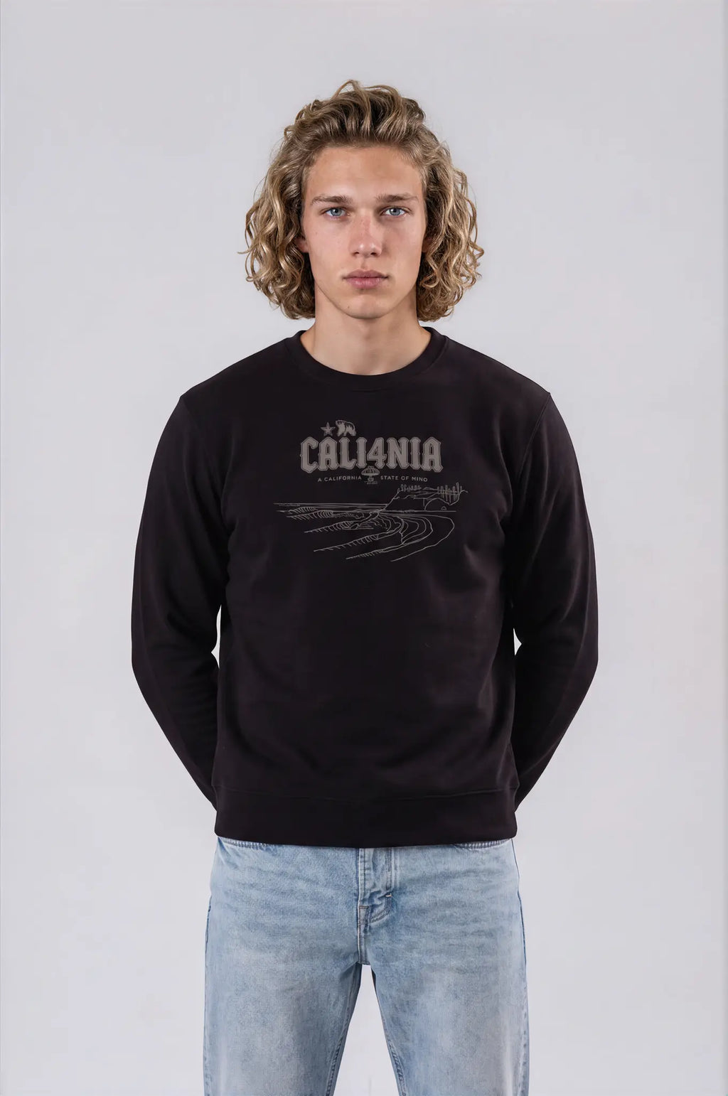 A young man with curly blonde hair wearing a black crewneck sweatshirt with a distressed white 'CALI4NIA' graphic on the chest, paired with light blue jeans, standing against a light gray studio background.