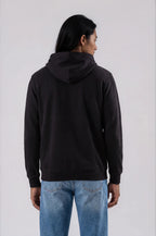 A man viewed from the back wearing a black pullover hoodie and blue jeans against a plain light gray studio background.